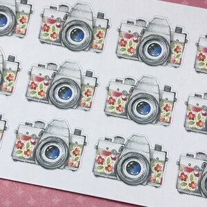 Pu&ograve; includere: Un foglio di adesivi con fotocamere in stile acquerello. Le fotocamere hanno un corpo argentato, una lente blu e sono decorate con fiori rossi e gialli. Gli adesivi sono su sfondo bianco.