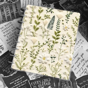 May include: A white notebook with a floral design embroidered on the cover. The notebook has a spiral binding and is surrounded by black and white paper scraps with inspirational quotes. The quotes include "Every child is gifted" and "Be full of joy".