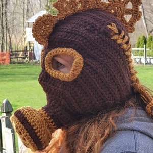 May include: A brown and tan crocheted balaclava-style hat with steampunk-inspired details. The hat features a circular eye opening, a snout-like projection, and decorative gear-shaped accents. The hat is made of a thick yarn and has a textured appearance.