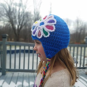 May include: A blue crocheted hat with a colorful flower design on the front. The hat has a long, braided cord that hangs down the back.
