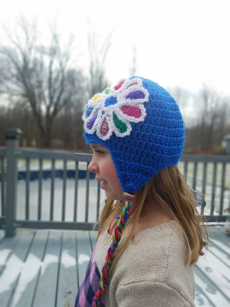 May include: A blue crocheted hat with a colorful flower design on the front. The hat has a long, braided cord that hangs down the back.