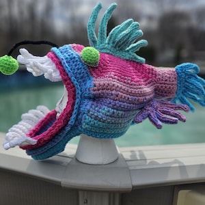Peut inclure: Un chapeau en crochet en forme de poisson-pêcheur avec un dégradé de rose, bleu et violet. Le chapeau a un leurre en crochet blanc et deux perles vertes pour les yeux.