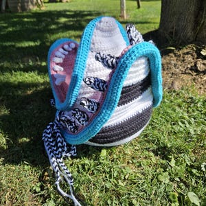 Könnte beinhalten: Ein handgefertigter Häkelhut mit einem einzigartigen Design. Der Hut hat eine blaue Krempe, einen rosa Innenraum mit weißen Zähnen und einen schwarz-weiß gestreiften Körper. Er hat schwarz-weiße geflochtene Bänder.