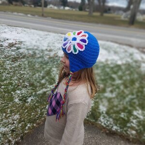 May include: A blue crocheted hat with a colorful flower design on top. The hat has ear flaps and a long, colorful braided tassel.
