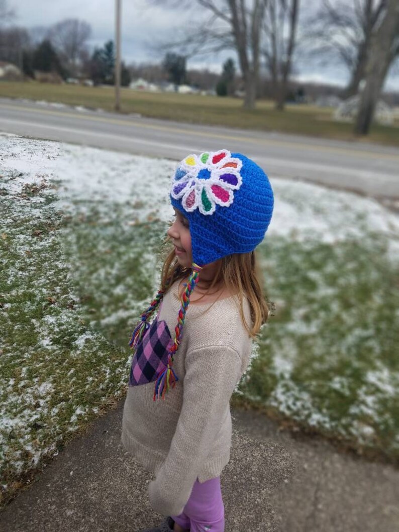 May include: A blue crocheted hat with a colorful flower design on top. The hat has ear flaps and a long, colorful braided tassel.