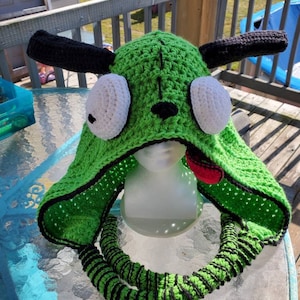 May include: A handmade green crochet hat with black accents, featuring large white eyes, black ears, and a red tongue. The hat has a neck warmer with black stripes. The hat is displayed on a mannequin head.