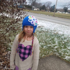 May include: A young person wearing a blue crocheted hat with a colorful flower on top. The hat has long colorful braids attached. The person is wearing a white sweater with a pink and black plaid heart on the front. The person is wearing purple pants.
