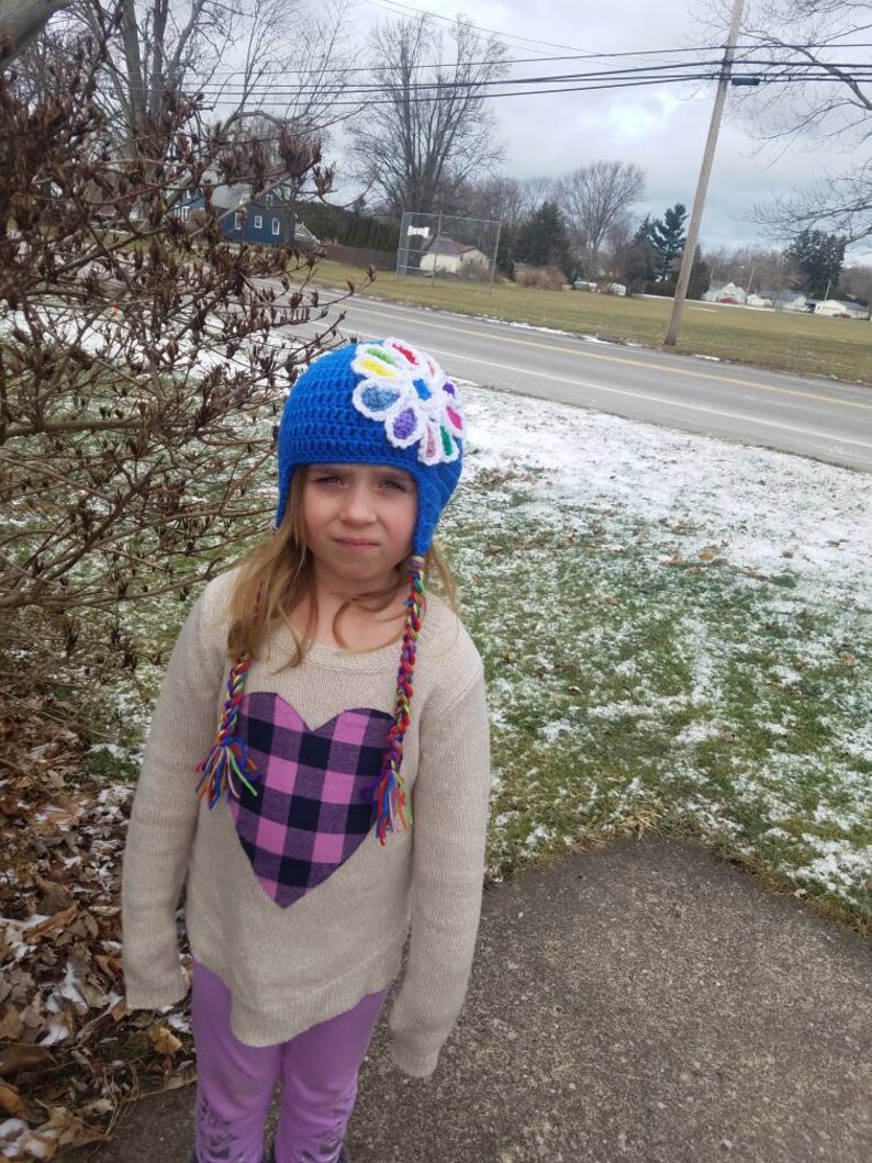 May include: A young person wearing a blue crocheted hat with a colorful flower on top. The hat has long colorful braids attached. The person is wearing a white sweater with a pink and black plaid heart on the front. The person is wearing purple pants.