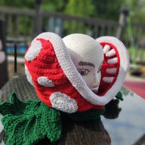 Puede incluir: Un sombrero de ganchillo hecho a mano que se asemeja a una planta Piraña de la serie de videojuegos Super Mario. El sombrero presenta una cabeza roja con manchas blancas, un borde blanco y una hoja verde. El sombrero se exhibe en una cabeza de maniquí.