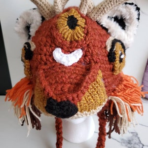 May include: A crocheted deer hat with a red, brown, and gold color scheme. The hat has a large, single eye with a black pupil and a white mouth. The hat has a long, brown cord that can be tied under the chin.