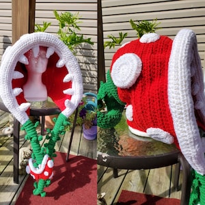 Pode incluir: Dois chapéus de crochê inspirados no jogo de vídeo Super Mario Bros. Um chapéu é vermelho e branco e se assemelha a um cogumelo. O outro chapéu é verde e branco e se assemelha a uma planta Piranha.