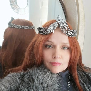 May include: A silver-colored crown with wolf head accents, worn over red hair. The crown is reflected in a mirror. The person is wearing a gray fur-trimmed garment and a silver necklace with a large ring pendant.