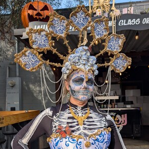 May include: A person in a Day of the Dead costume with elaborate skull face paint, a decorative headpiece, and a corset with blue and white floral patterns. The costume includes skeleton detailing and a butterfly accent.