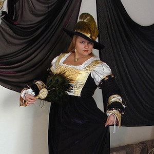 May include: A woman in a Renaissance-style costume. The black dress has gold and white accents on the bodice and sleeves. She wears a black hat with gold feathers and holds a peacock feather fan. The costume is a full-length gown.