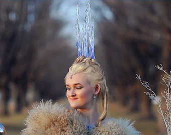 May include: A woman in a light blue dress and a fluffy beige shawl holds a silver sphere and a bundle of silver branches. She wears a jeweled headpiece and a blue and silver crown. The background is a blurred outdoor scene.