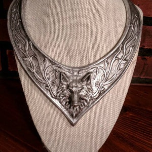 May include: A silver-colored statement necklace featuring a detailed wolf head pendant. The necklace has an intricate design with floral and vine-like patterns. It is displayed on a beige linen bust against a brick background.
