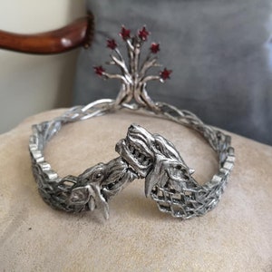 May include: A silver crown with a tree design and red embellishments. The crown has a braided band and two wolf heads facing each other. The crown is displayed on a beige cushion.