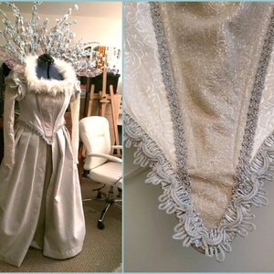 May include: A pale, floor-length gown with a decorative bodice and sleeves. The dress features silver trim and a faux fur collar. A matching headpiece with floral accents completes the ensemble. The dress is displayed on a mannequin.