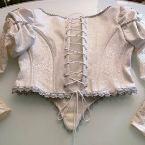 May include: Ivory corset top with long sleeves. The bodice has a floral pattern and a lace-up back. Silver trim accents the neckline, sleeves, and hem. The sleeves have a puffed shoulder design. A unique garment for special occasions.