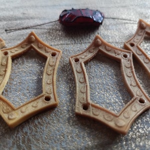 May include: Close-up of several tan diamond-shaped frames with decorative edges and small holes. A dark red, irregularly shaped bead is suspended above the frames. The frames have a vintage or antique aesthetic.