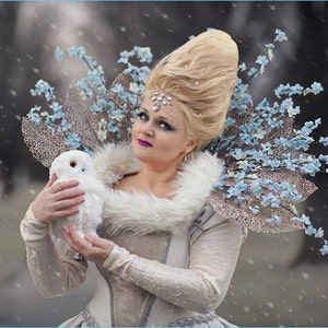 May include: A woman in a winter-themed costume holds a white owl. She wears a white dress with a fur collar, a decorative headpiece, and a floral shoulder piece. The background is a blurred outdoor scene with falling snow.