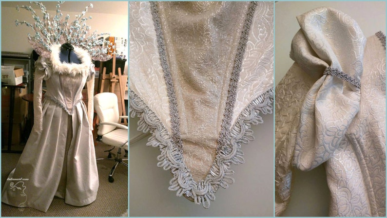 May include: A pale, floor-length gown with a decorative bodice and sleeves. The dress features silver trim and a faux fur collar. A matching headpiece with floral accents completes the ensemble. The dress is displayed on a mannequin.