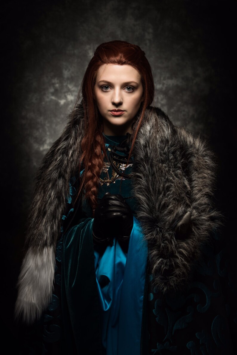May include: A portrait of a person with long red hair, wearing a teal dress and a faux fur stole. The person is holding black gloves. The background is a dark, textured gray.