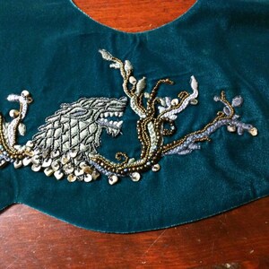 May include: A dark teal velvet fabric with an embroidered design. The embroidery features a wolf-like creature with intricate detailing, surrounded by beaded branches and gold sequins. The design is centered on the fabric.