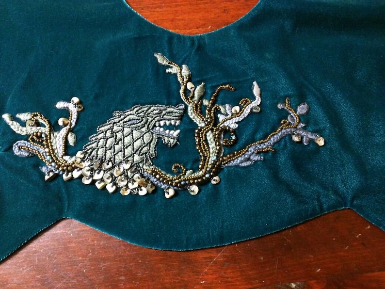 May include: A dark teal velvet fabric with an embroidered design. The embroidery features a wolf-like creature with intricate detailing, surrounded by beaded branches and gold sequins. The design is centered on the fabric.