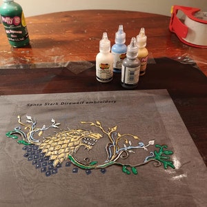 May include: An embroidery project featuring a direwolf design with the text "Sansa Stark Direwolf embroidery." The design uses gold, blue, green, and white paint. Several bottles of paint in various colors are visible, along with a roll of tape.