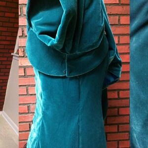 May include: A teal velvet gown with long sleeves and a hood, displayed on a mannequin. The dress features a high collar with embroidered detailing and flared sleeves with blue satin cuffs. The back of the dress has a corset-style lace-up closure.