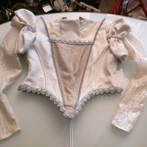 May include: An ivory-coloured bodice with long sleeves. The garment has a floral pattern, puffed sleeves, and silver trim. The bodice has a V-shaped front panel and a high neckline. The sleeves are long and fitted at the wrist.