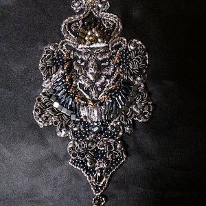 Puede incluir: Un broche plateado ornamentado con un diseño de corona. El broche presenta intrincados abalorios en tonos negro, plateado y dorado. El diseño es simétrico, con una base en forma de lágrima y una corona detallada en la parte superior.