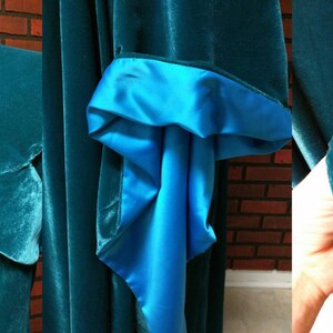 May include: Teal velvet cloak with a contrasting bright blue satin lining. The cloak features a high collar with decorative clasps and a laced back. The sleeves have a flared design with a satin cuff and black lace trim. A medieval-inspired garment.