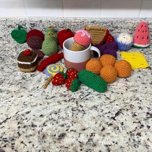 May include: A selection of crocheted food items in various colours, including an ice cream cone, cupcake, strawberries, and oranges. Other items include a pear, apple, and watermelon slice. The items are arranged on a speckled countertop.
