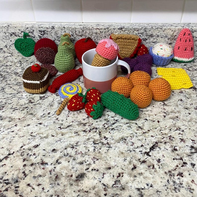 May include: A selection of crocheted food items in various colours, including an ice cream cone, cupcake, strawberries, and oranges. Other items include a pear, apple, and watermelon slice. The items are arranged on a speckled countertop.