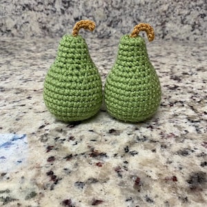 May include: Two handmade crochet pears. The pears are green with a brown stem. The pears are on a speckled countertop.