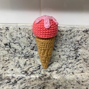 May include: A crocheted ice cream cone. The ice cream is a bright pink with light pink drizzles, sitting on a golden-brown cone. The cone has a textured appearance, and the item is made from yarn.