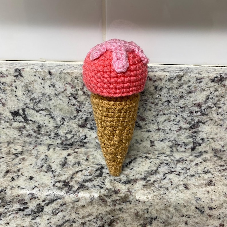May include: A crocheted ice cream cone. The ice cream is a bright pink with light pink drizzles, sitting on a golden-brown cone. The cone has a textured appearance, and the item is made from yarn.