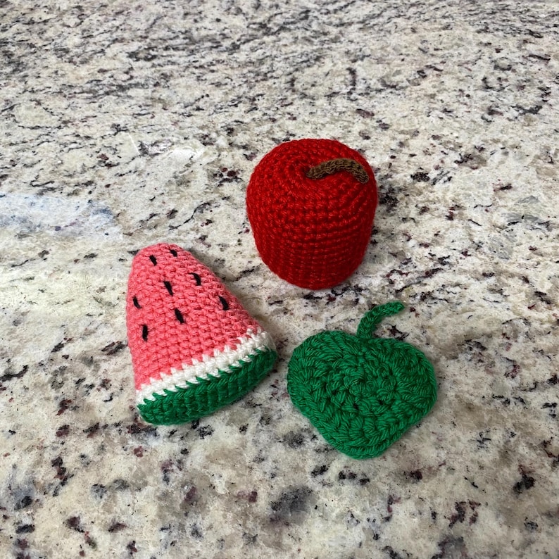 May include: Three crocheted food items are displayed: a red apple with a brown stem, a watermelon slice with black seeds, and a green leaf. The items are handmade and rest on a speckled surface.