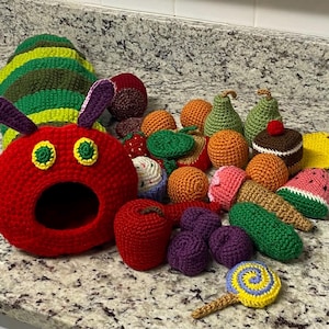 May include: A selection of handmade crochet items, featuring a red caterpillar with a large opening, a striped caterpillar, and various food items such as oranges, pears, and a slice of watermelon. The items are in various colours and textures.