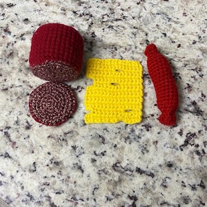 May include: A selection of handmade crochet food items, including a red burger bun, a burger patty, a slice of yellow cheese, and a red hot dog. The items are arranged on a speckled surface.