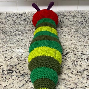 May include: A handmade, crocheted caterpillar toy. The caterpillar has a red head with purple antennae, and a body composed of alternating green, yellow, and brown sections. The toy is designed to resemble a caterpillar.