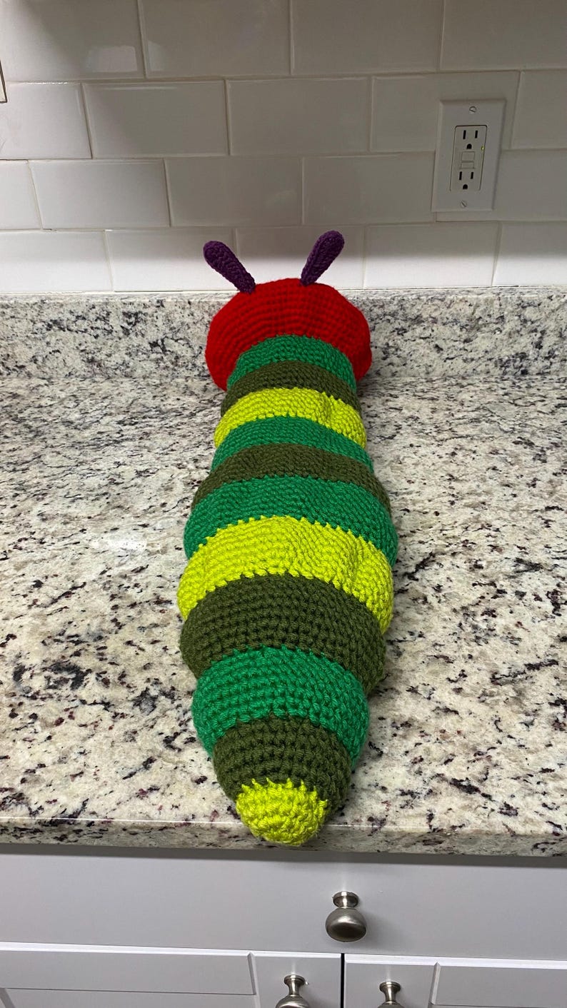 May include: A handmade, crocheted caterpillar toy. The caterpillar has a red head with purple antennae, and a body composed of alternating green, yellow, and brown sections. The toy is designed to resemble a caterpillar.
