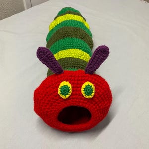 May include: A colourful, handmade crochet caterpillar toy. The head is red with yellow and green eyes and purple antennae. The body is striped with green, yellow, and olive green sections. The caterpillar has an open mouth.