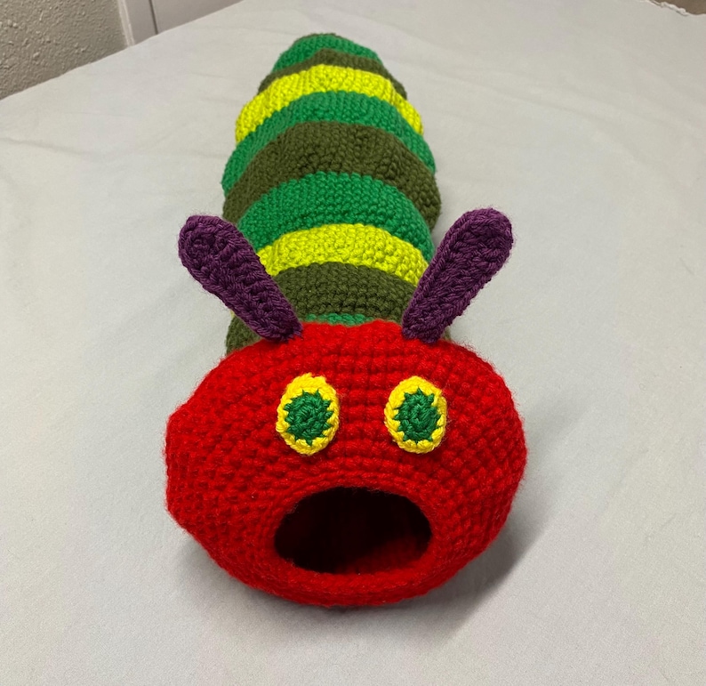 May include: A colourful, handmade crochet caterpillar toy. The head is red with yellow and green eyes and purple antennae. The body is striped with green, yellow, and olive green sections. The caterpillar has an open mouth.