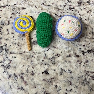 May include: Three crocheted food items are displayed: a lollipop with a yellow and blue spiral, a green pickle, and a cupcake with white frosting and colourful sprinkles. All items are handmade.