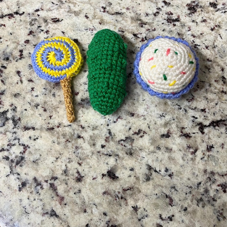 May include: Three crocheted food items are displayed: a lollipop with a yellow and blue spiral, a green pickle, and a cupcake with white frosting and colourful sprinkles. All items are handmade.