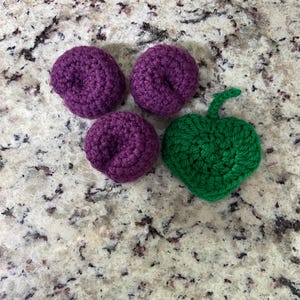 May include: Four crocheted fruit shapes on a speckled surface. Three are purple, resembling plums, and one is green, shaped like an apple. These handmade items are likely for decoration or children's play.