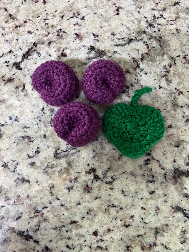 May include: Four crocheted fruit shapes on a speckled surface. Three are purple, resembling plums, and one is green, shaped like an apple. These handmade items are likely for decoration or children's play.